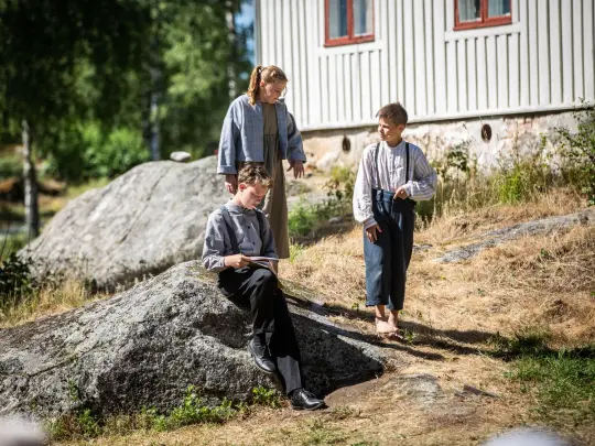 gutt som sitter og tegner, to som står og ser på